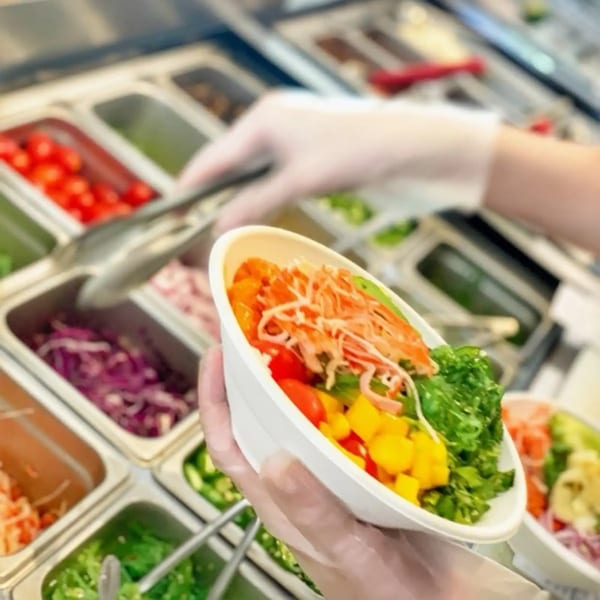 Gloved hands preparing a colorful poke bowl with fresh ingredients from a serving bar.