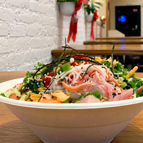 A colorful poke bowl with crab, tuna, salmon, noodles, and greens on a wooden table.