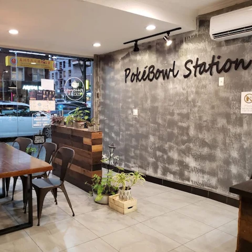 Interior of PokiBowl Station with a wooden table, chairs, plants, and a textured gray wall.