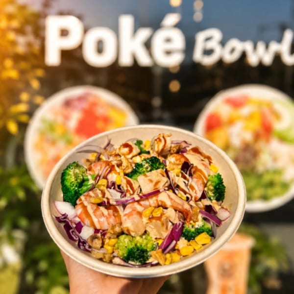 A hand holds a poke bowl with shrimp, broccoli, corn, and red cabbage, in front of a "Poké Bowl" sign.