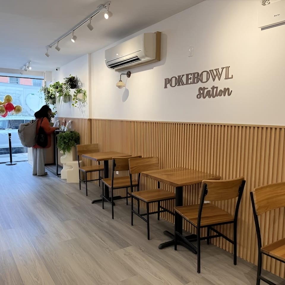 Interior of Pokebowl Station with wooden tables and chairs, light wood paneling, and a customer at the counter.