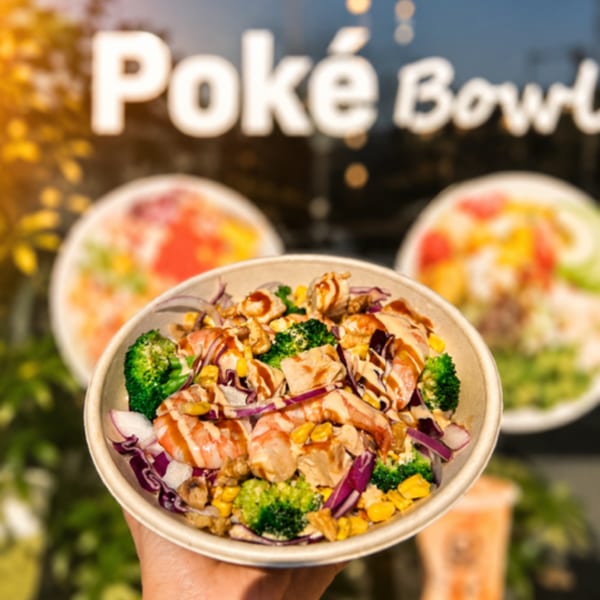 A hand holds a poke bowl with shrimp, chicken, broccoli, corn, and red onion.
